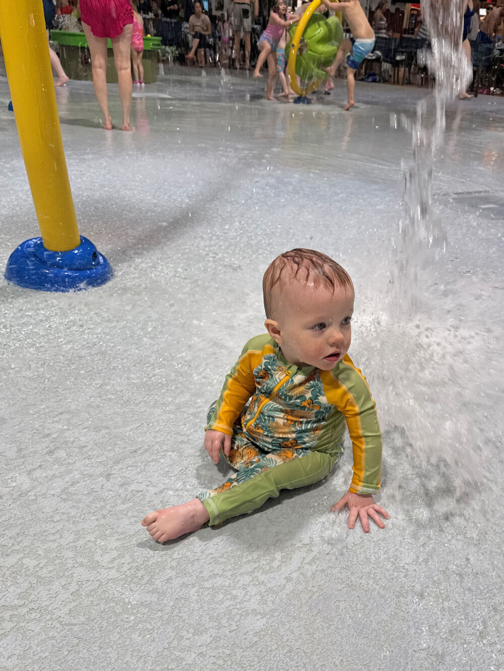 A baby wearing a full-coverage green rash guard sitting and splashing in a shallow waterpark area.