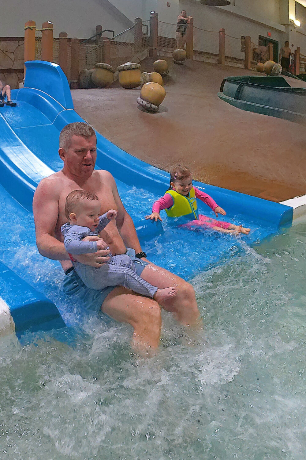 A father sliding down a toddler-friendly blue waterslide while holding his baby son, with his daughter sliding next to them.