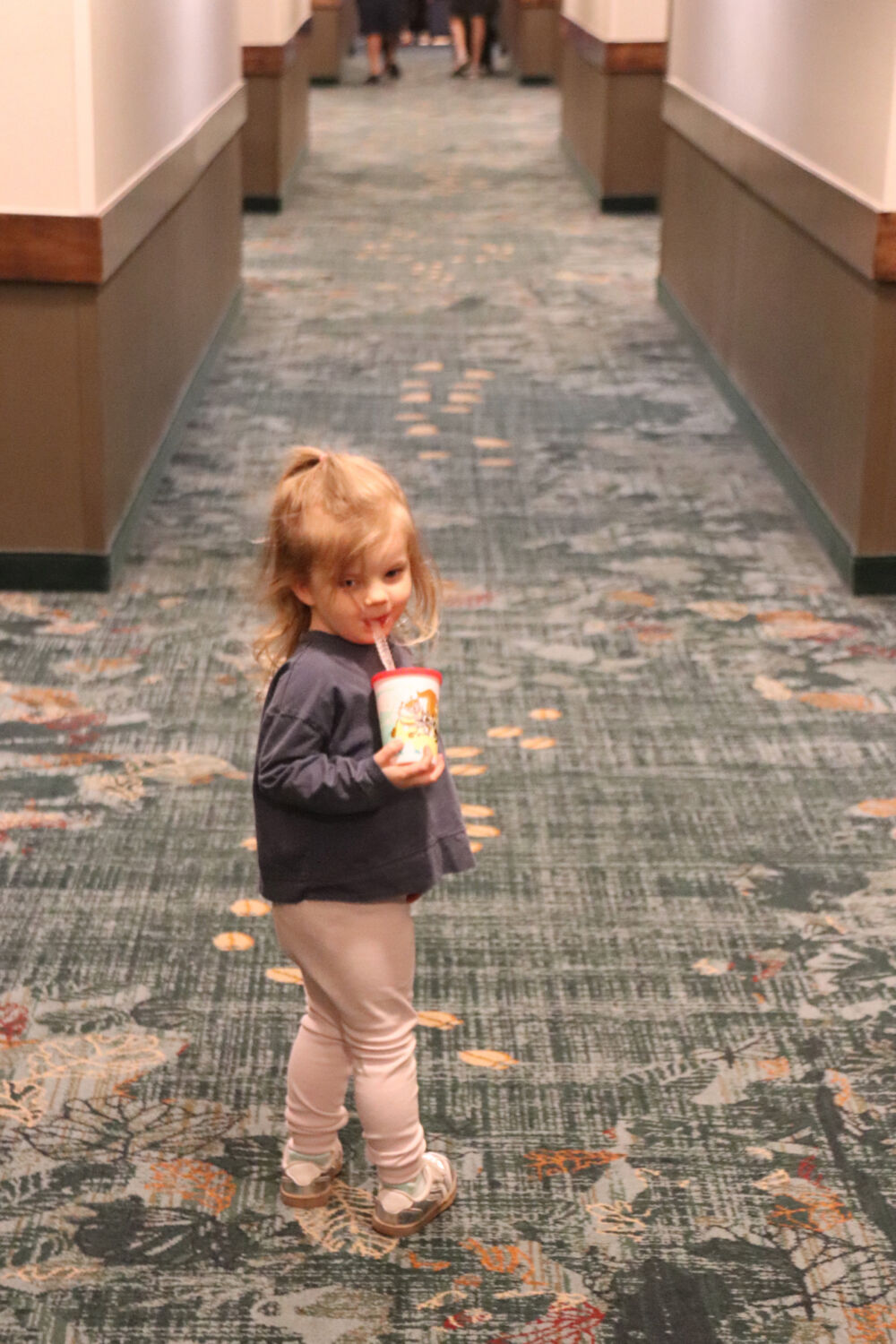 A toddler girl standing in a long resort hallway holding a character cup.