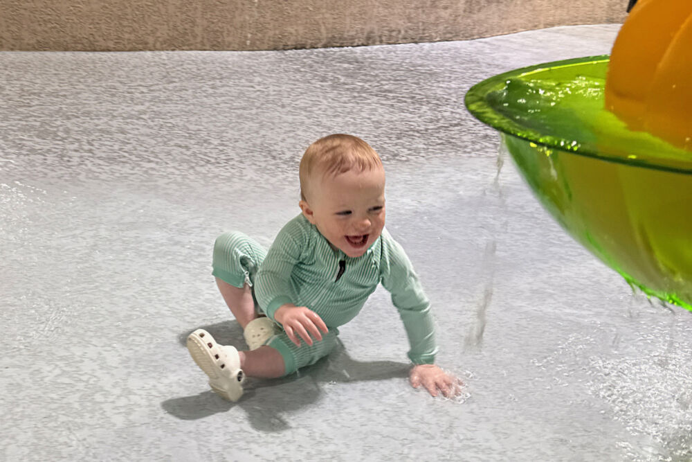 A baby crawling and laughing in the shallow water of a splash zone.