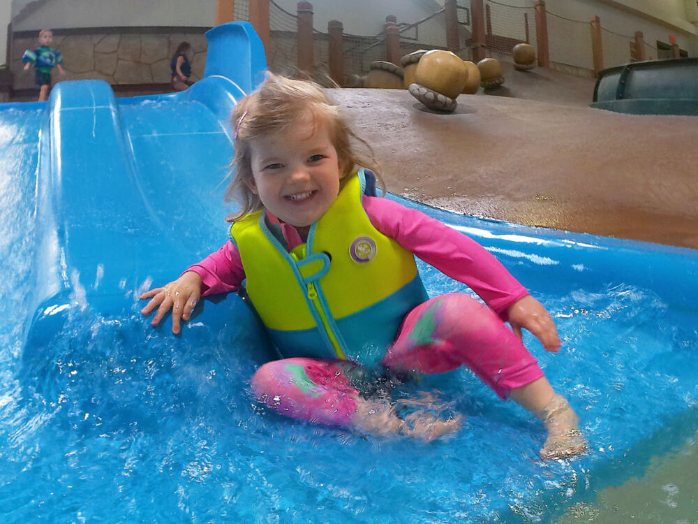 A young girl in a yellow and blue life jacket and pink swimsuit smiling as she lands in the shallow water at the bottom of a blue waterslide.
