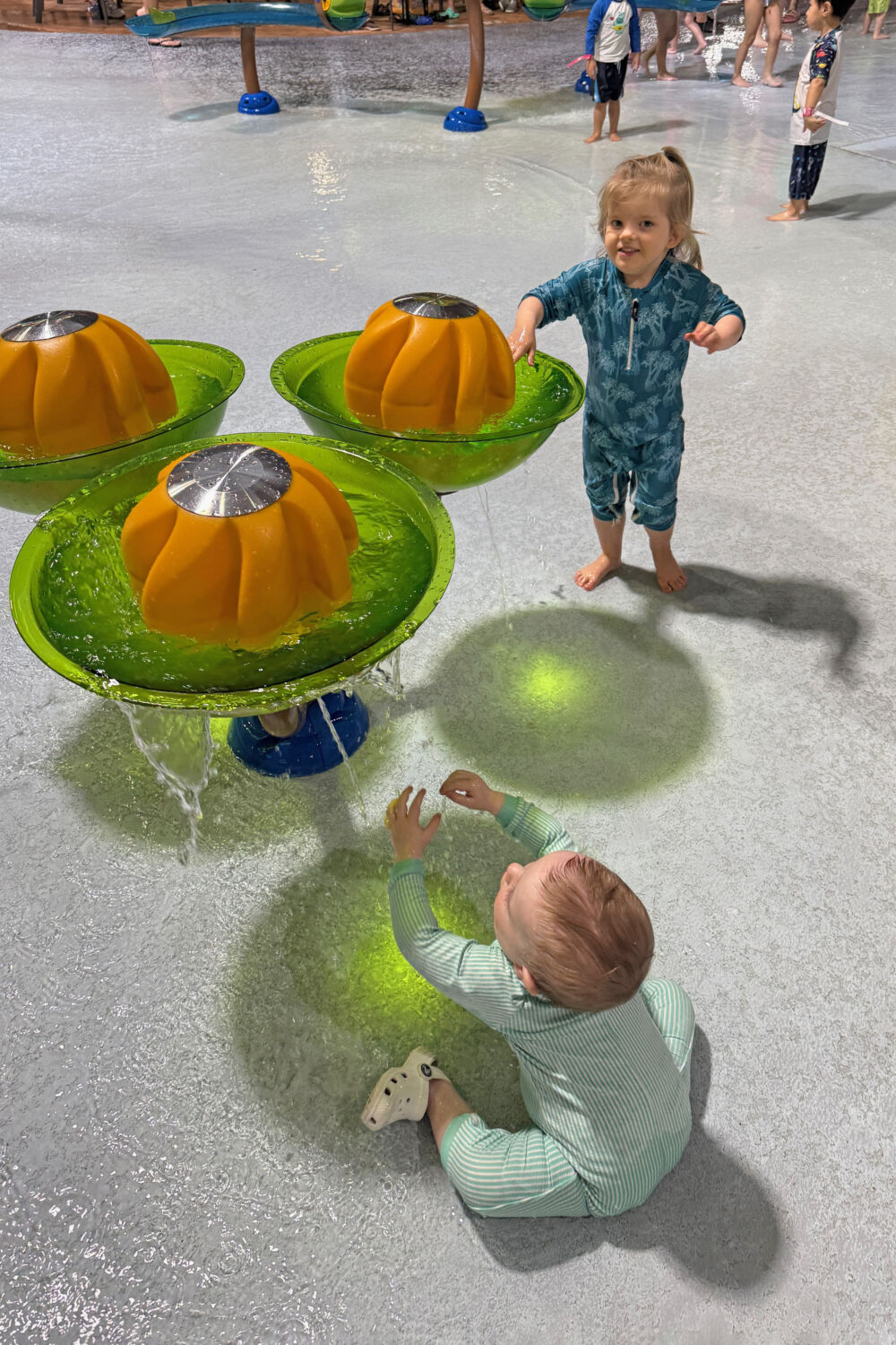 Two young children playing with interactive orange and green water fountain features on a splash pad.