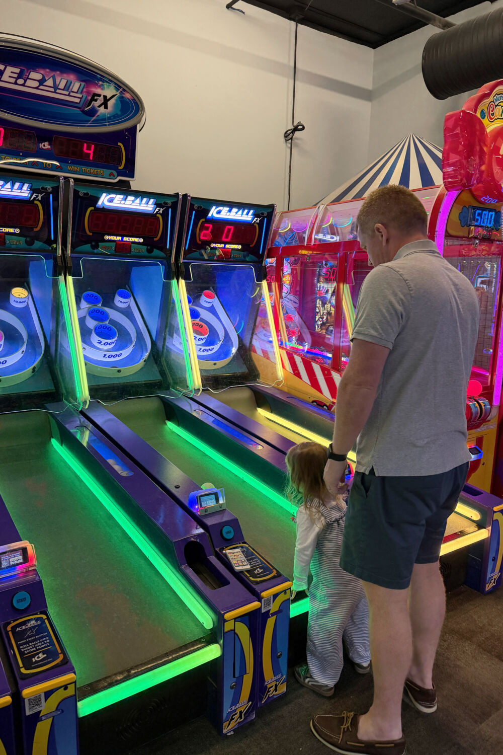 A father helping his daughter roll balls up the ramp of a Skee-Ball machine.