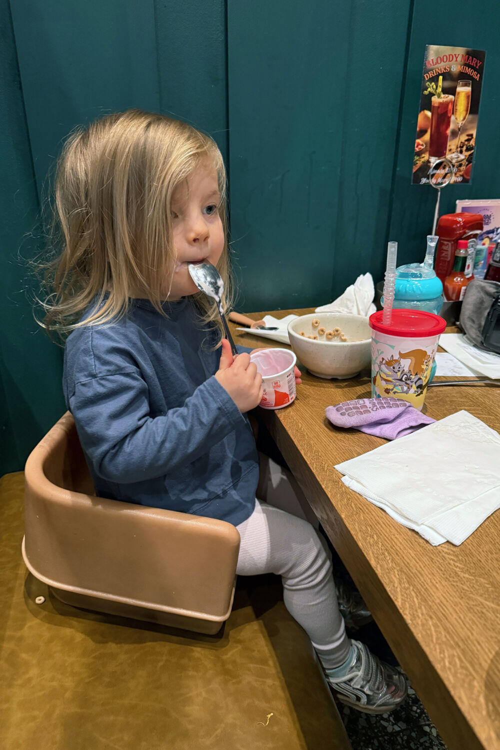 A toddler girl sitting in a booster seat at a dining table, enjoying her meal and using a straw cup.