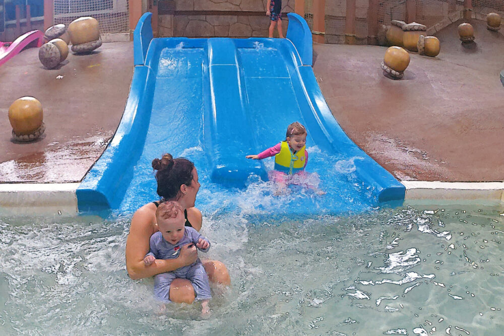 A mother holding a baby and a toddler girl sliding down a small blue waterslide into a shallow pool.